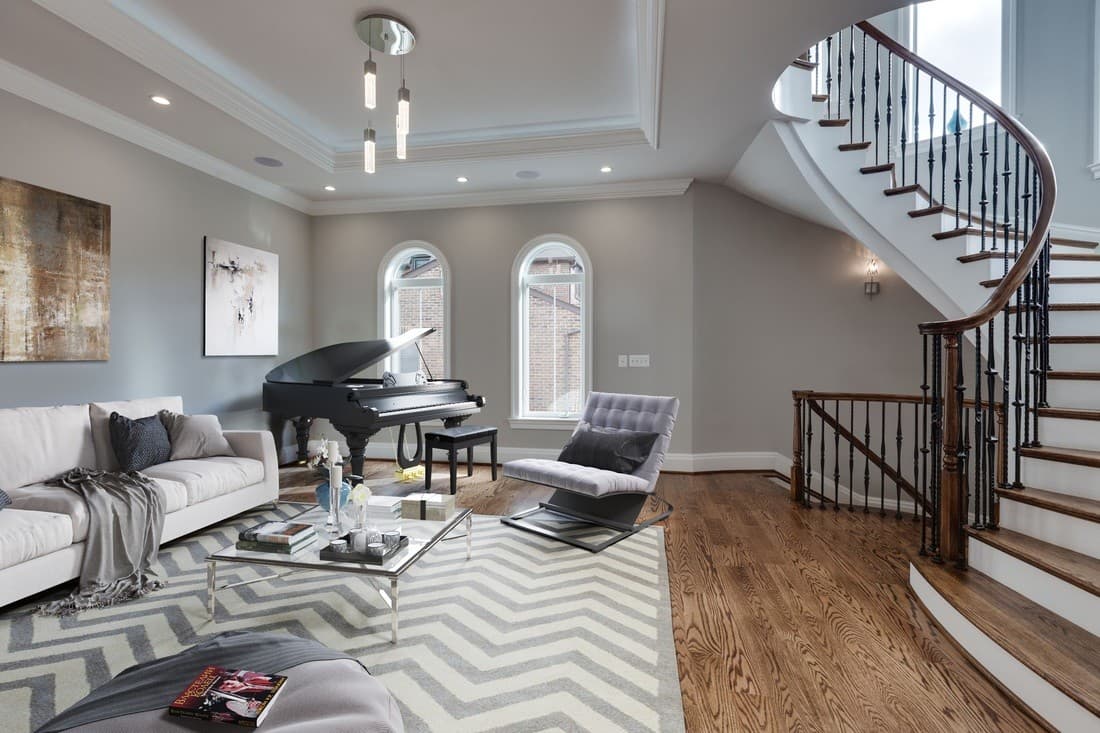 The same room virtually staged with a grand piano, sofa, lounge chairs and a patterned rug beside the curved staircase.